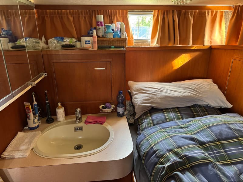 Een kajuit met een bed met geruit dekbed, een wastafel met kraan en spiegelkast, en diverse toiletartikelen.