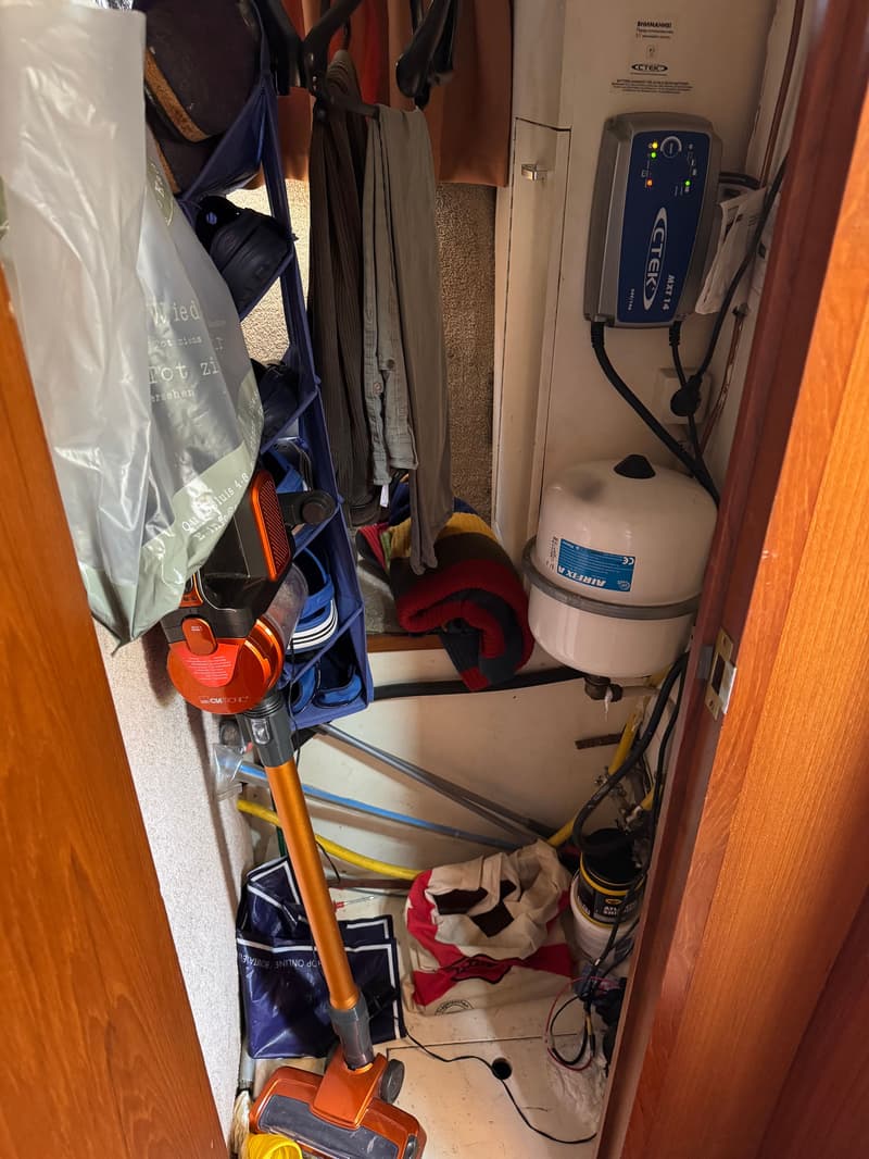 Een opbergruimte met een stofzuiger, kleding, schoenen, een acculader en een expansievat.