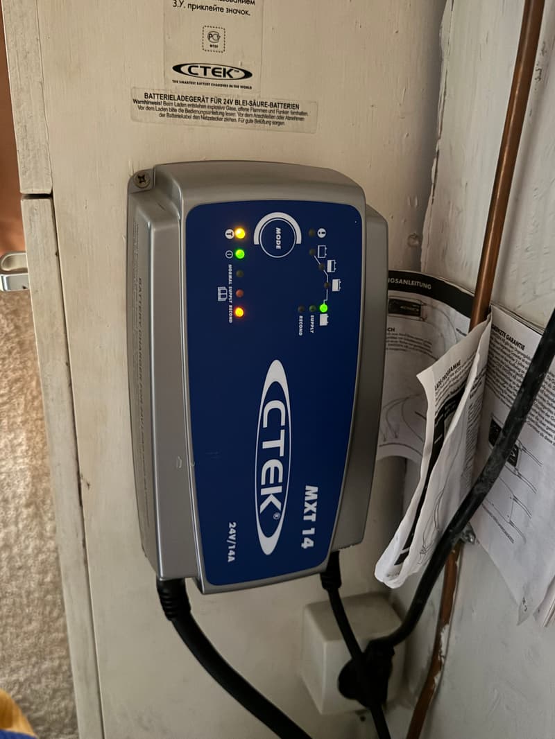 Een CTEK MXT 14 acculader met verlichte indicatoren is gemonteerd op een lichte wand, met kabels en handleidingen ernaast.