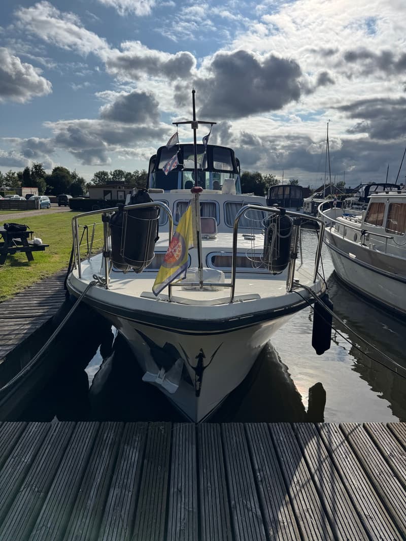 Een witte motorboot ligt aangemeerd aan een houten steiger, met een gele vlag aan de boeg en stootwillen aan de zijkanten.
