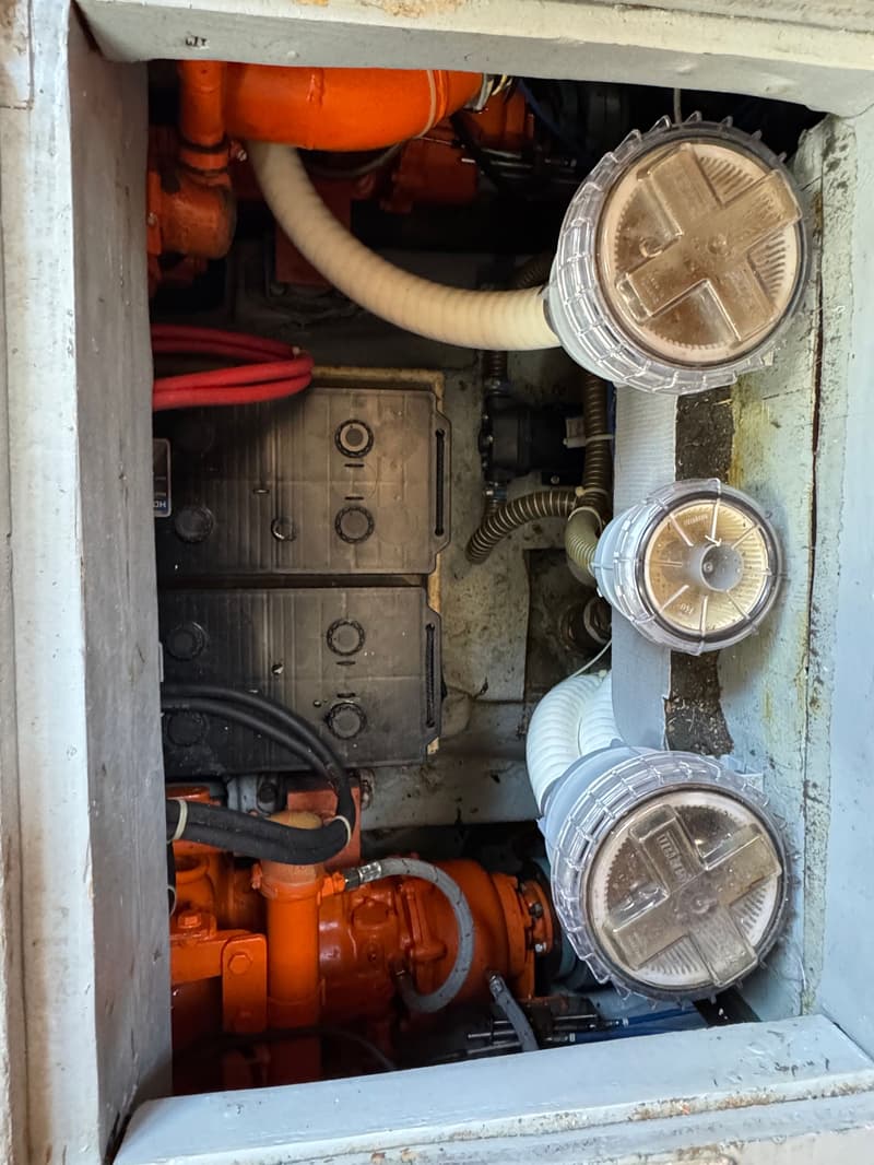 De foto toont een close-up van een motorruimte met oranje motoronderdelen, accu's en drie waterfilters.