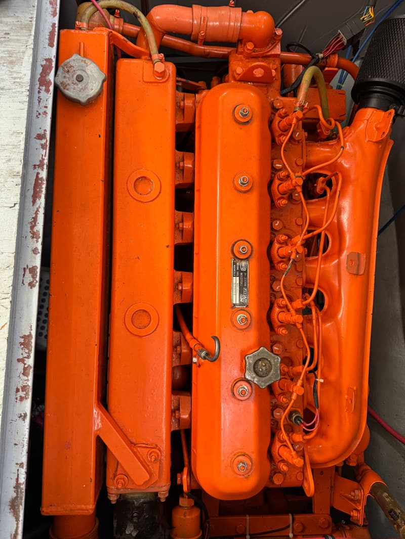 Een close-up van een oranje scheepsmotor met diverse leidingen, bouten en een metalen plaatje met tekst.