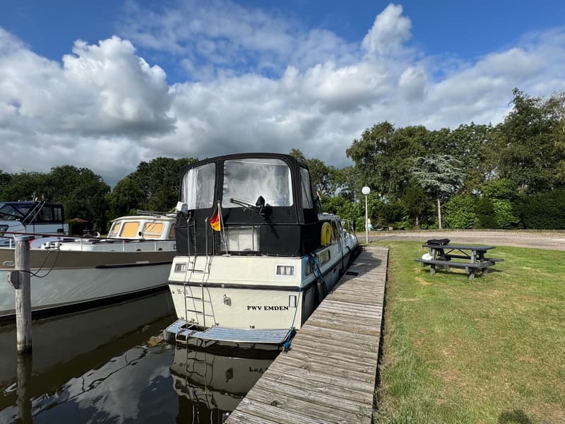 Een witte boot met een zwarte kap en de tekst "PWV EMDEN" op de spiegel ligt aangemeerd aan een houten steiger.
