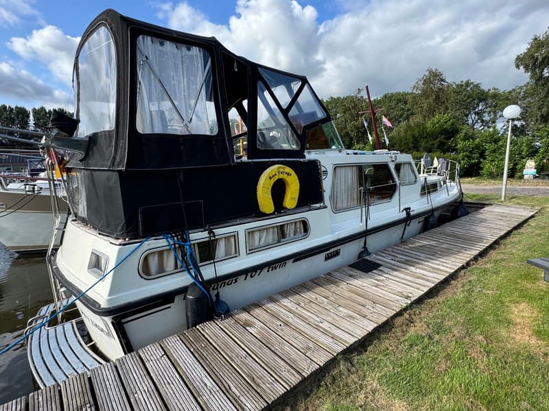 Een witte motorboot met zwarte kap en gele reddingsboei ligt aangemeerd aan een houten steiger. — Neptunus 107 TWIN