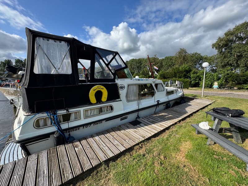 Een witte motorboot met zwarte kap en gele reddingsboei ligt aangemeerd aan een houten steiger. — Neptunus 107 TWIN