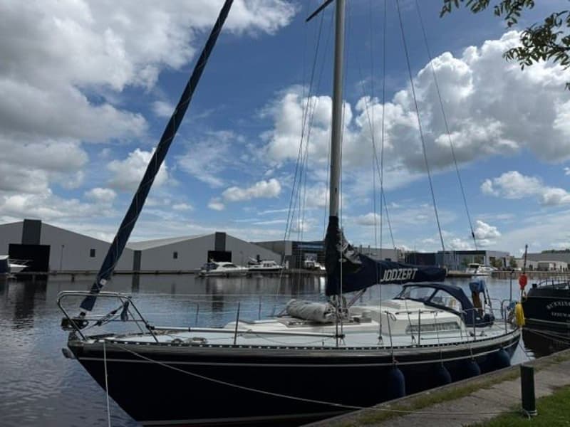 Een donkerblauwe zeilboot met een witte opbouw ligt aangemeerd aan een kade, met een blauwe zeilhoes met de naam "JODZERT".