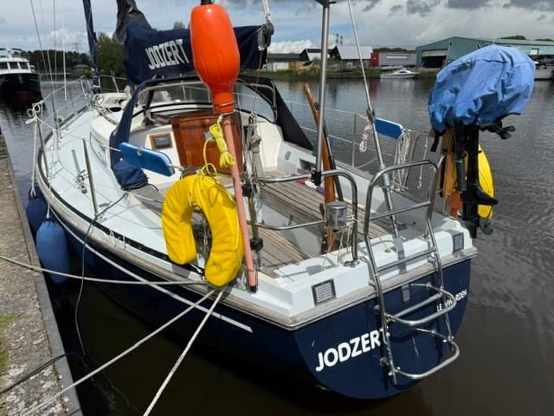 Een blauw-witte zeilboot genaamd "JODZERT" ligt afgemeerd aan een kade, met een oranje boei en gele reddingsboei.