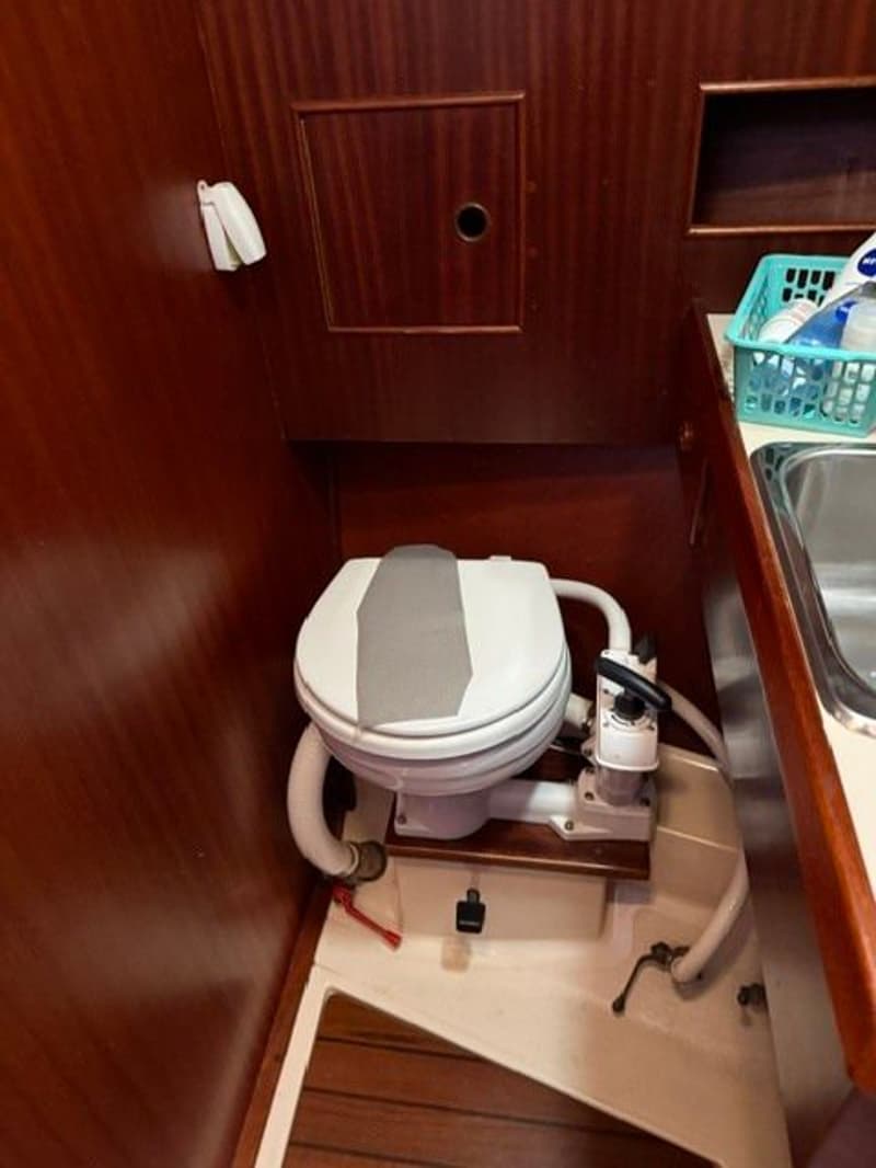 Een boottoilet met een grijze zitting en een handpomp, omringd door houten wanden en een gootsteen.