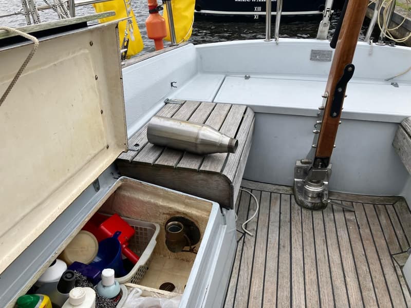 Een open opbergvak met diverse schoonmaakartikelen en een metalen fles op een houten bankje in de kuip van een boot.