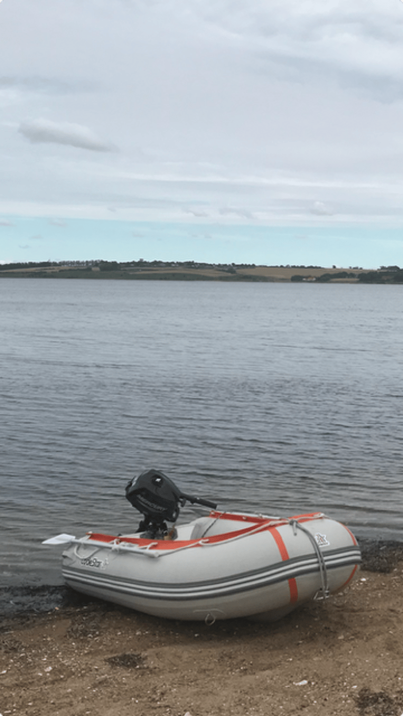 Een grijze opblaasboot met een zwarte buitenboordmotor en oranje accenten ligt op een kiezelstrand bij water.