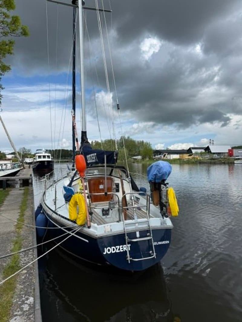 Een blauwe zeilboot met de naam "JODZERT" ligt aangemeerd aan een kade onder een bewolkte hemel. — Victoire 34 S-spant