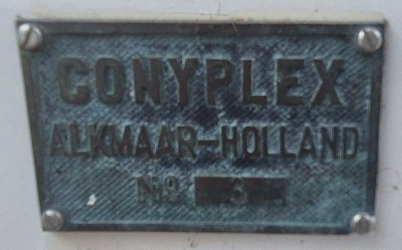 Een metalen plaatje met de tekst "CONYPLEX ALKMAAR-HOLLAND NO 5", vastgeschroefd met vier schroeven.