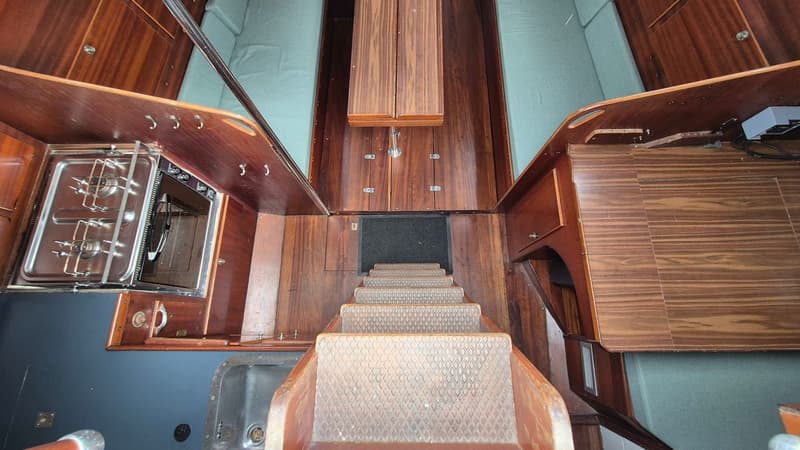 De foto toont een houten kajuitinterieur met een trap, een fornuis met oven, een gootsteen en zitbanken met groene kussens.
