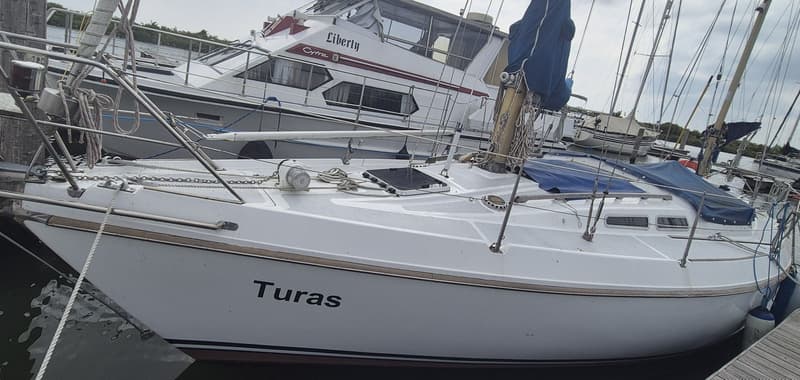 Een witte zeilboot met de naam "Turas" ligt afgemeerd naast een steiger, met een andere boot op de achtergrond.