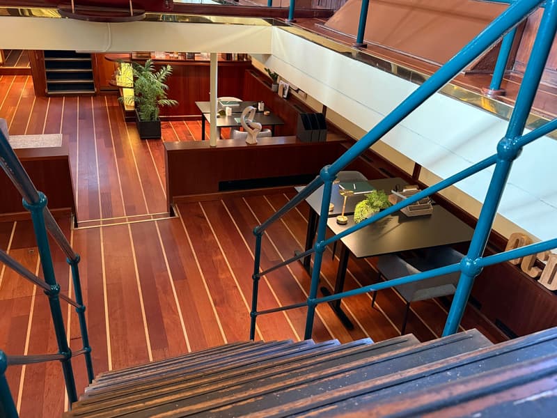 Een houten trap met blauwe reling leidt naar een lager dek met houten vloer, tafels, stoelen en planten.