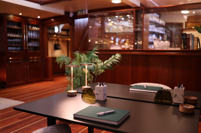 Een interieur met een donkere tafel, twee lampen, notitieboekjes en pennen, omringd door houten wanden en kasten.
