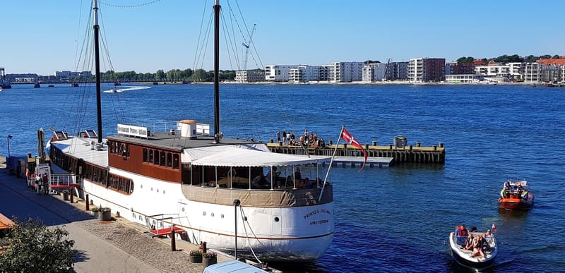 Een grote witte boot met bruine opbouw en twee masten ligt aangemeerd, met "PRINSES JULIANA AMSTERDAM" op de spiegel.