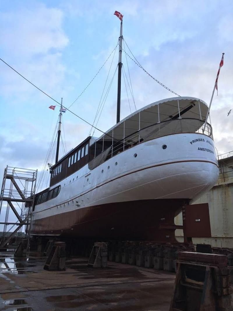 Een witte en bruine boot, genaamd "PRINSES JULIANA AMSTERDAM", staat op houten blokken in een droogdok onder een bewolkte…