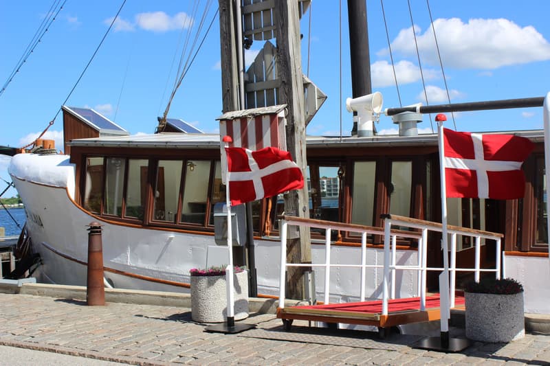 Een witte boot met bruine ramen ligt aangemeerd aan een kade met kasseien, met twee Deense vlaggen en een loopplank.