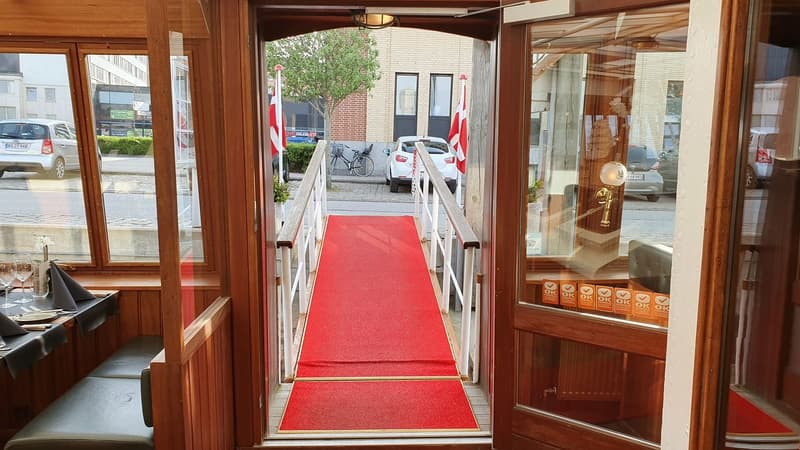 Een rode loper leidt vanaf een houten interieur met tafels en banken naar buiten, geflankeerd door twee Deense vlaggen.
