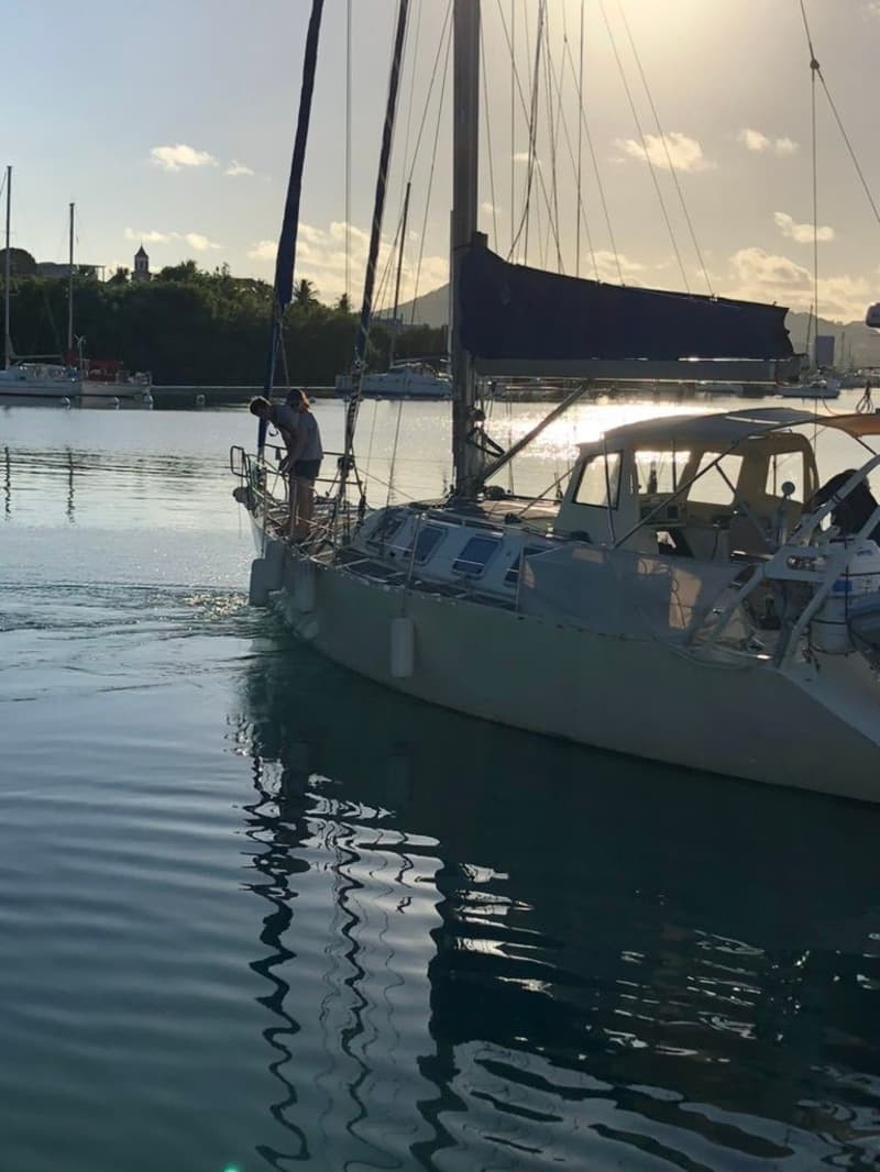 Een zeilboot met een persoon op de boeg ligt in het water, met een deels opgerold grootzeil en fenders aan de zijkant.