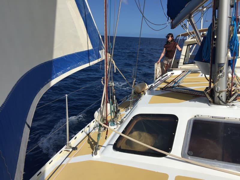 Een zeilboot vaart op open water met een deels uitgerold wit zeil met blauwe band, en een persoon op het dek.