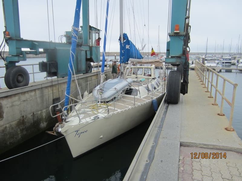 Een lichtgekleurde zeilboot met de naam "Tepee" op de boeg ligt in het water naast een hijskraan in een haven.