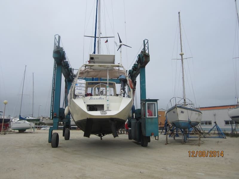 Een zeilboot met de naam "Bruxelles" op de spiegel wordt door een blauwe botenlift op het droge getild, met andere boten op…