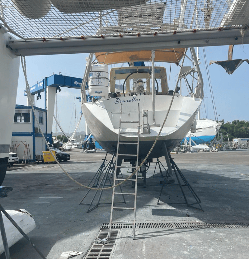 Een witte zeilboot met de naam "Bruxelles" op de spiegel staat op bokken op de wal, met een ladder ertegenaan.