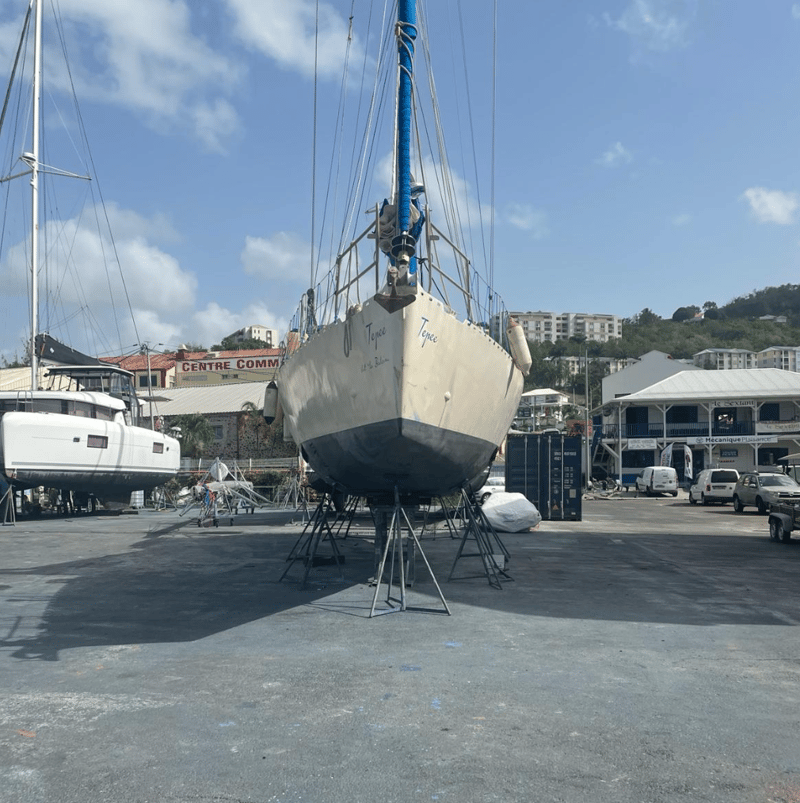 Een zeilboot genaamd 'Tepee' staat op bokken in een botenwerf, met een catamaran en gebouwen op de achtergrond.