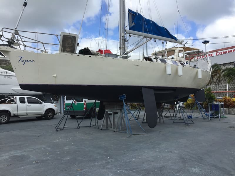 Een lichtgekleurde zeilboot met de naam "Tepee" op de boeg staat op bokken op de wal, met een opgerold blauw zeil.