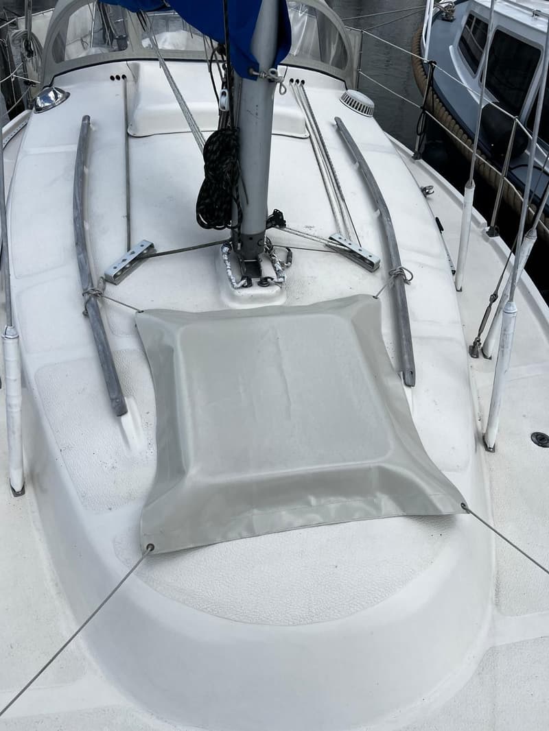 Een wit scheepsdek met antislipstructuur, een grijze rechthoekige hoes, een mast met blauw zeil en diverse lijnen zijn…