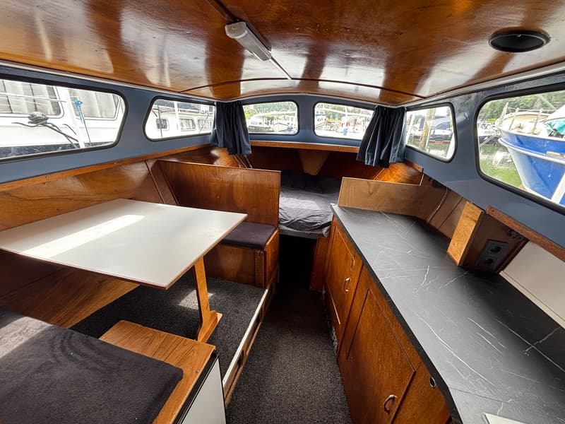 De foto toont het interieur van een boot met houten wanden en plafond, een tafel, een bed en een aanrechtblad.