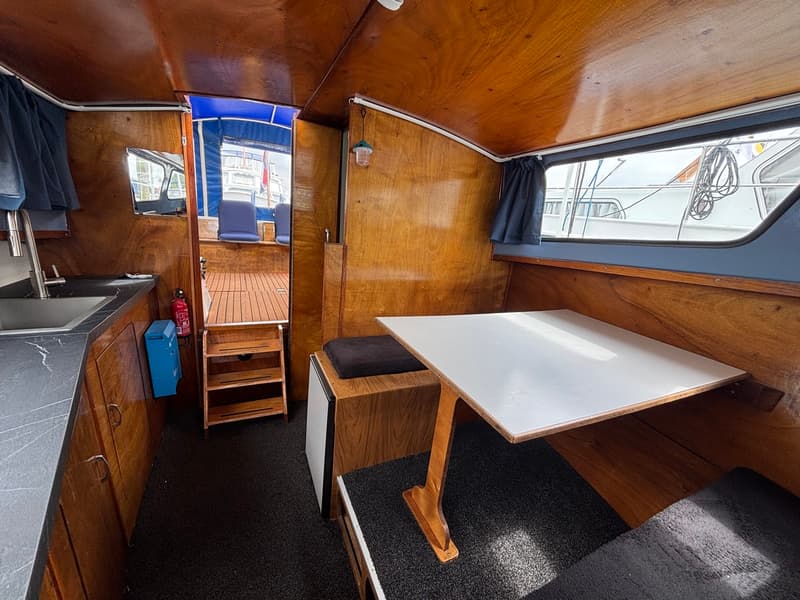 De foto toont het interieur van een boot met houten wanden, een keukenblok, een tafel met banken en een doorgang naar buiten.