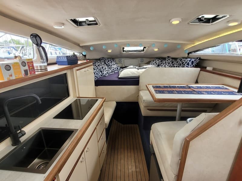 Een interieur van een boot met een keukenblok, zithoek en bed, verlicht door plafondlampen en daglicht.