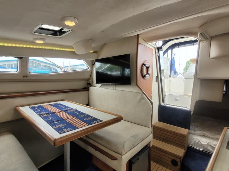 De foto toont het interieur van een boot met een zithoek, een tafel, een televisie en een doorgang naar een ander gedeelte.