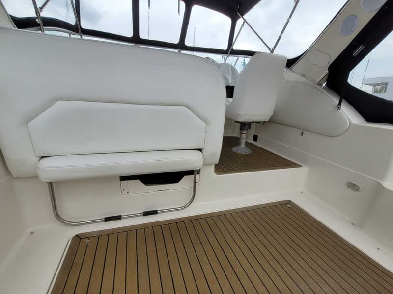 De foto toont een bootinterieur met witte gestoffeerde zitplaatsen, een bruine houten vloer en een stuurstoel.