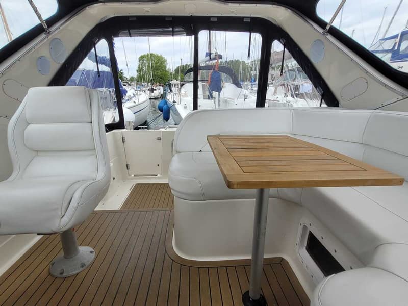 De foto toont het interieur van een boot met witte zitbanken, een houten tafel en een uitzicht op een jachthaven.