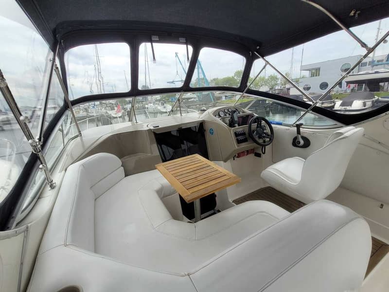 De foto toont het interieur van een boot met witte zitbanken, een houten tafel en een stuurwiel.