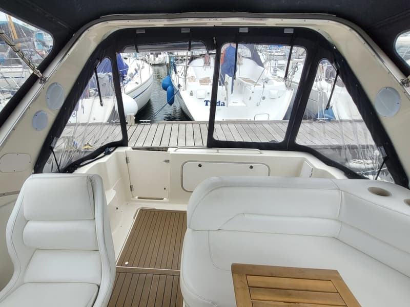 De foto toont het interieur van een boot met witte stoelen en banken, een houten vloer en een uitzicht op andere boten in…