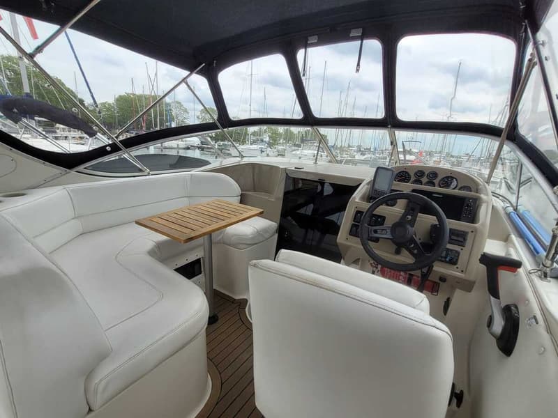 Een bootinterieur met witte banken, een houten tafel en een stuurconsole met instrumenten, met uitzicht op een jachthaven.