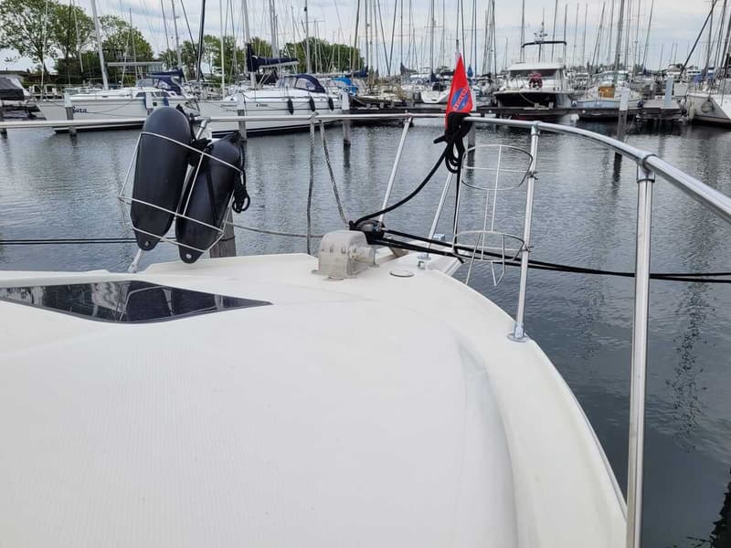 De foto toont de boeg van een witte boot met twee zwarte stootwillen in een houder, een rode vlag en een reling.