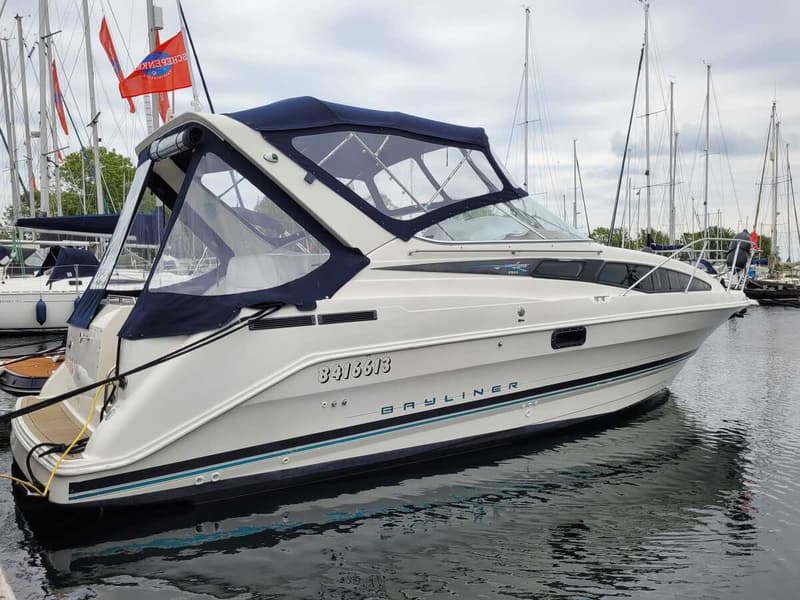 Een witte motorboot met een blauwe kap en ramen ligt in het water, met andere boten op de achtergrond.