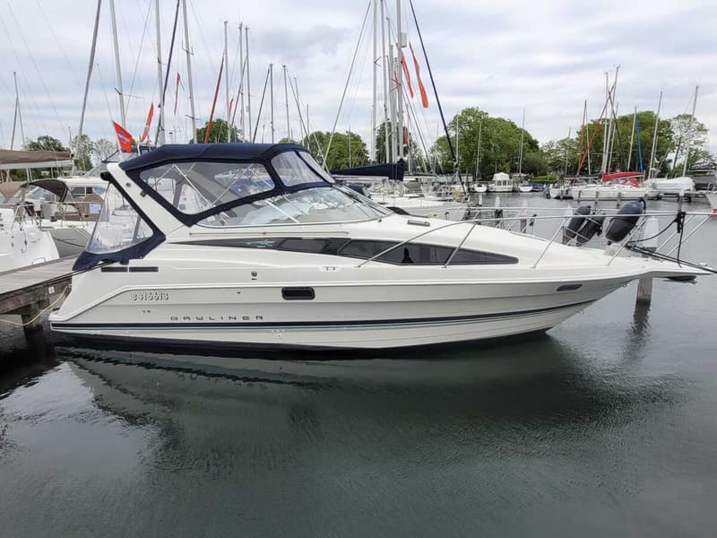 Een witte Bayliner motorboot met blauwe kap en strepen ligt aangemeerd aan een houten steiger in een haven.