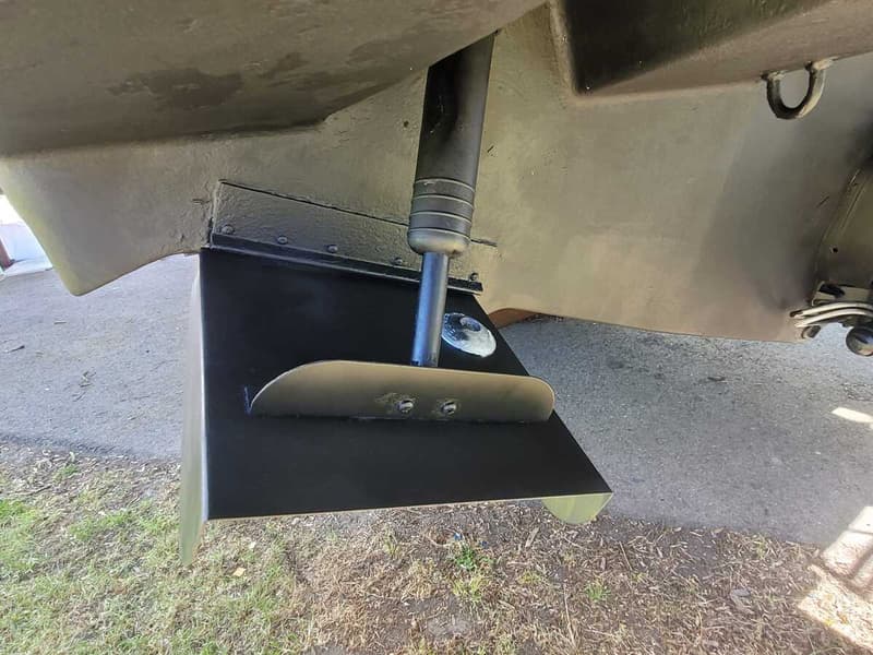 Een detailfoto van een zwart roer en een deel van de romp van een boot, met gras en bestrating op de achtergrond.