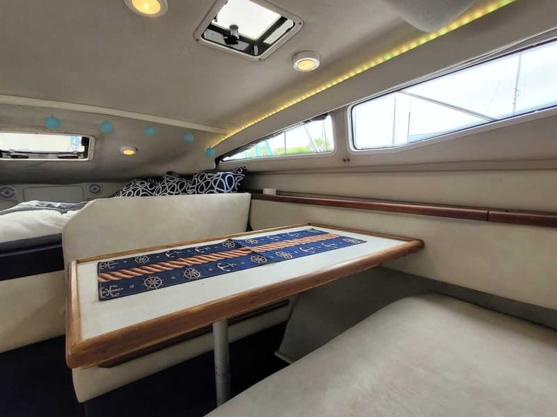 De foto toont het interieur van een boot met een tafel, banken, een bed, ramen en verlichting.