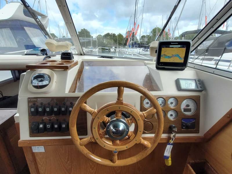 Een houten stuurwiel, een kompas, een Raymarine plotter en diverse meters en schakelaars zijn zichtbaar op het dashboard…