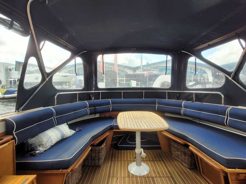 Een bootinterieur met een U-vormige blauwe bank met witte biezen, een houten tafel en een teakhouten vloer.