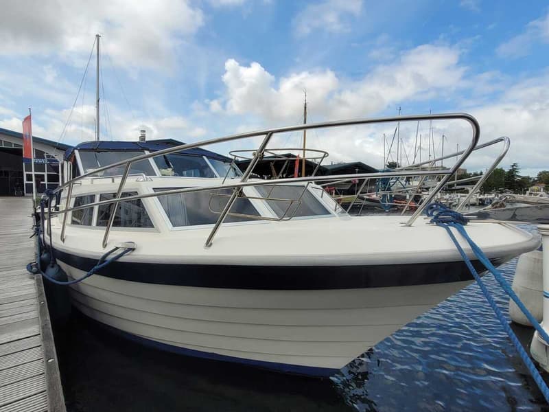 Een witte motorboot met een blauwe streep en een blauw dekzeil ligt afgemeerd aan een houten steiger. — Agder 840 OK