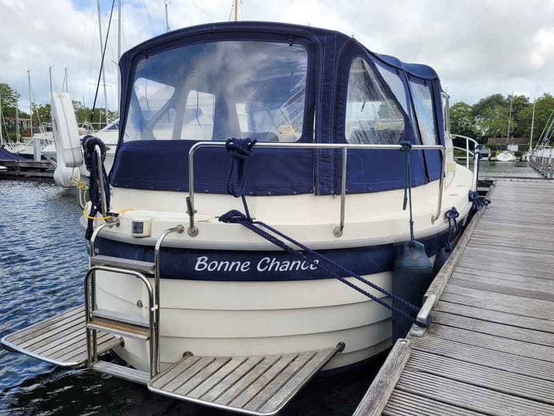 Een witte boot met blauwe accenten en een blauwe kap ligt afgemeerd aan een houten steiger. — Agder 840 OK